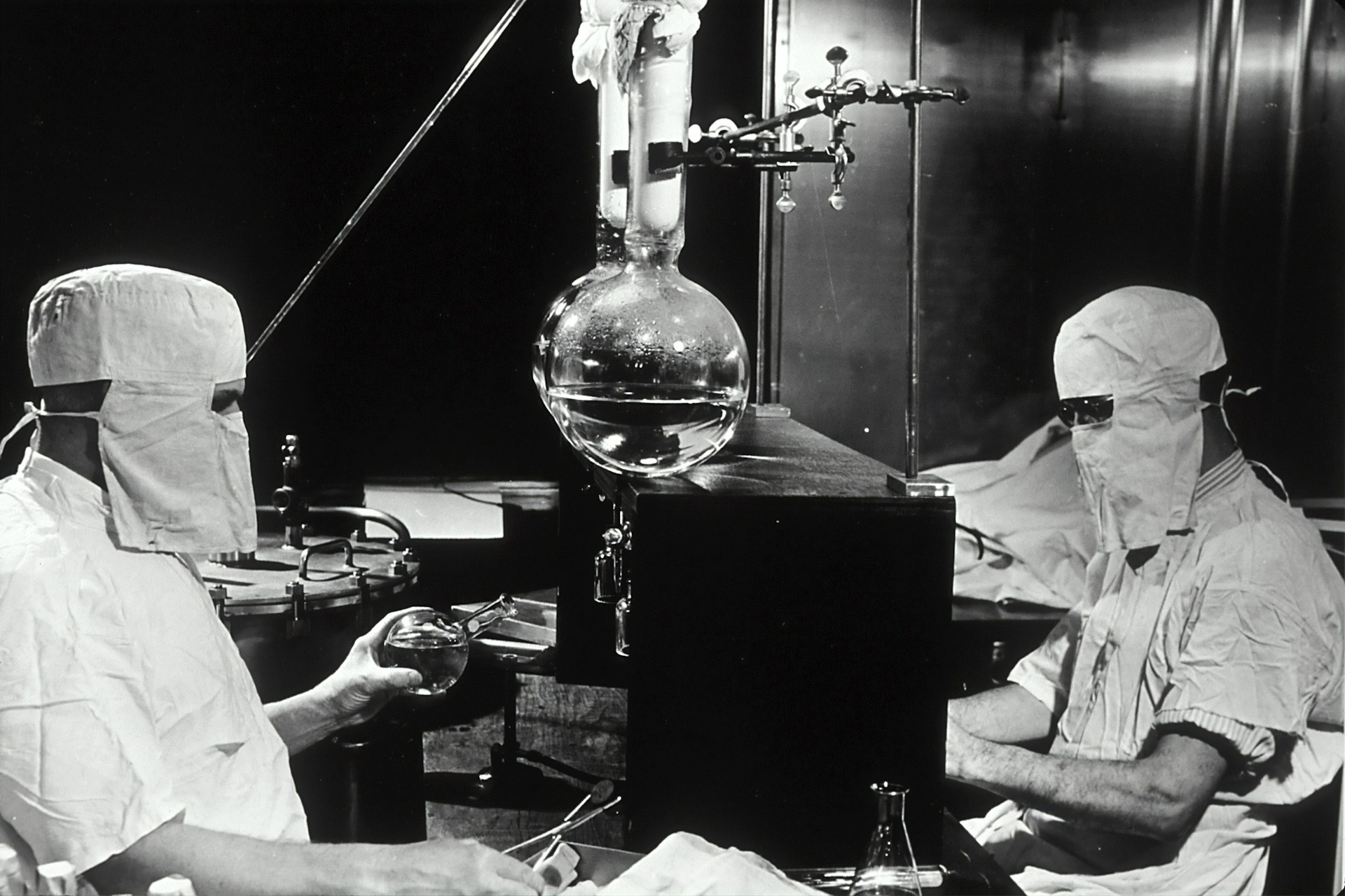 Archive photograph of two masked, hooded technicians working either side of a large distillation flask