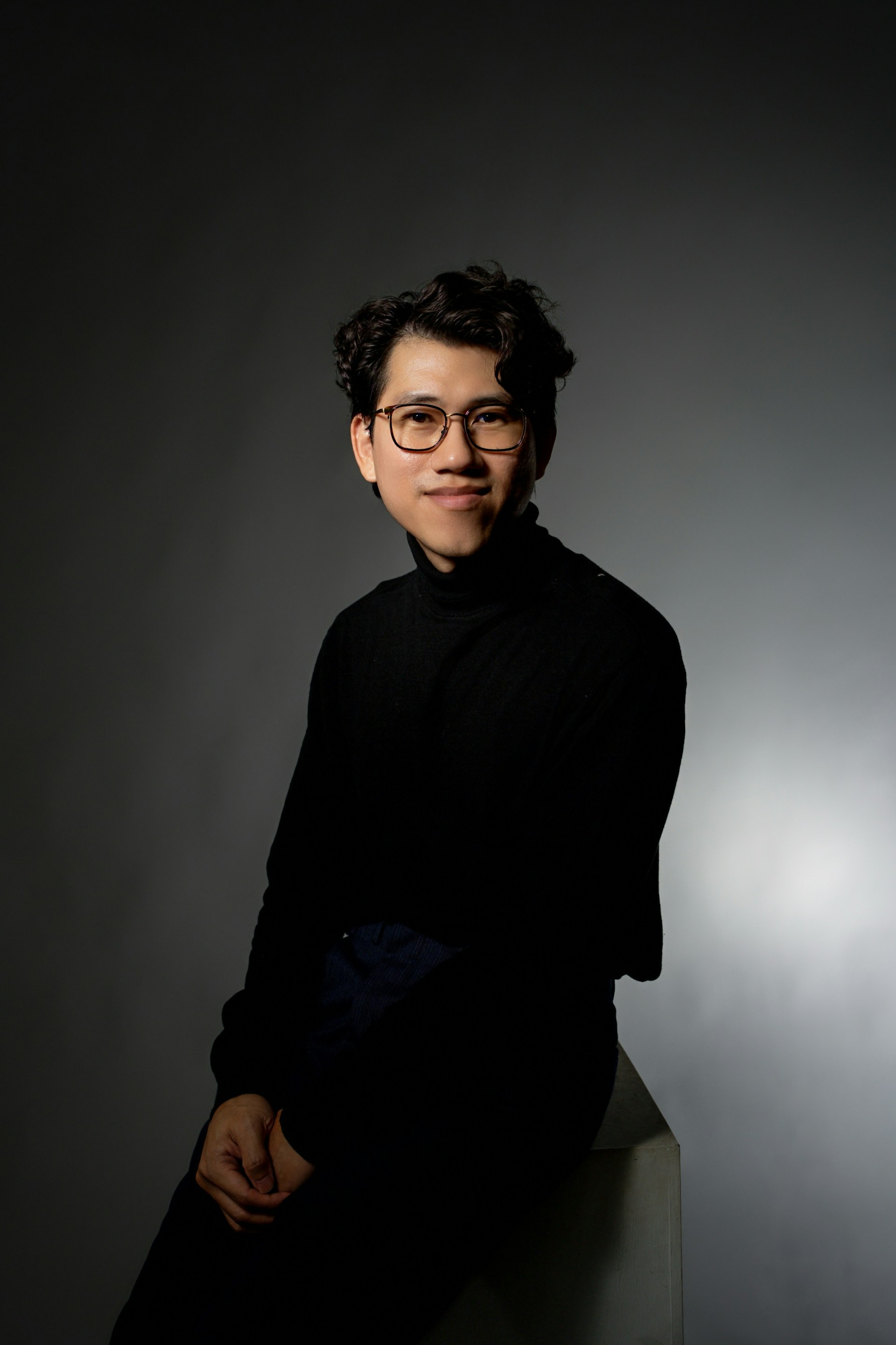 Portrait: a young man in glasses and a black turtleneck, seated against a grey backdrop